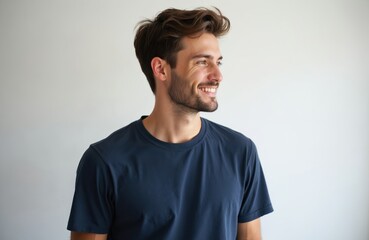Handsome young man poses against white background wearing blue t-shirt. Attractive male with modern hairstyle, beard smiles, looking side. Cheerful, happy person with natural face, confident