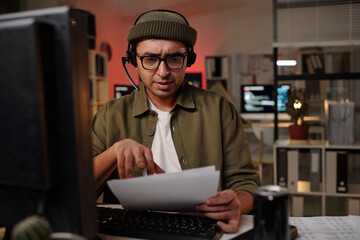 South Asian man wearing headset sitting at desk handling documents while working at computer in...