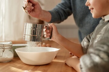 Senior man helping child sifting flour into bowl in kitchen, hands and partial faces visible,...