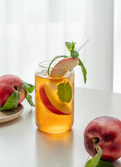Ice tea with peach, mint in a glass on a white table near window with fresh fruits and sunlight.
