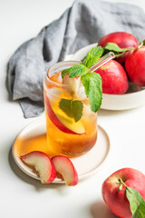 Ice tea with peach, mint in a glass on a white table near window with fresh fruits, napkin and sunlight.
