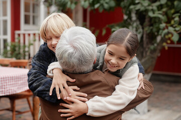 Senior Caucasian man embracing smiling boy and girl outdoors in village setting, children wrapping...