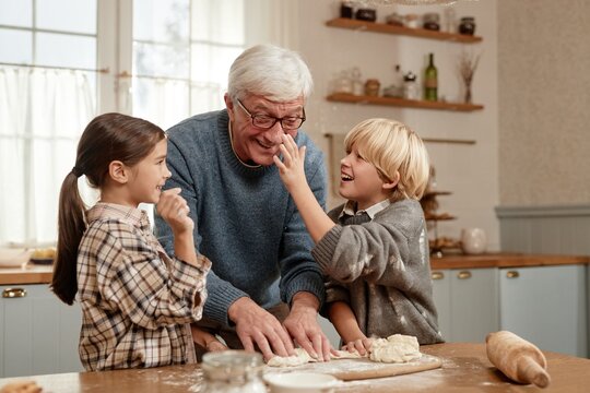 Senior Caucasian man smiling while preparing dough with two children in kitchen, girl and boy laughing and interacting with grandfather during baking activity in village home