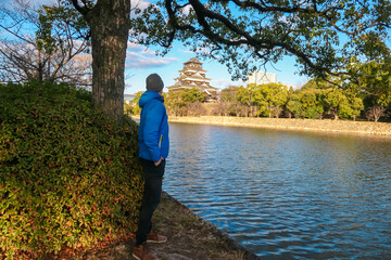 Male tourist in a blue jacket stands by the moat, admiring the view of iconic Hiroshima Castle. He enjoys a peaceful moment of contemplation while exploring the historic landmark in Hiroshima, Japan