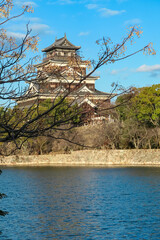 Historic Hiroshima Castle, known as the Carp Castle, rises majestically beyond its tranquil moat. This scenic view in Hiroshima, Japan, is framed by delicate tree branches against a clear blue sky