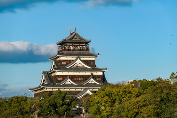 Magnificent keep of Hiroshima Castle rises above a lush canopy of trees. Traditional Japanese architecture set against brilliant blue sky with soft clouds, a perfect day at. Historic landmark in Japan