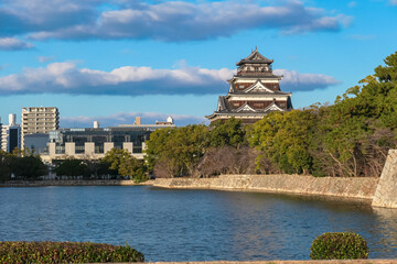 Magnificent Hiroshima Castle, a historic landmark in Japan, rises above its tranquil moat and lush trees. Scene juxtaposes ancient architecture with the modern city skyline of Hiroshima under blue sky