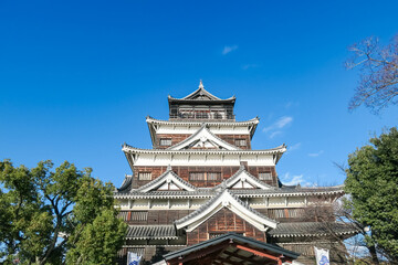 Magnificent main keep of Hiroshima Castle, a historic landmark in Japan, is beautifully framed by lush green trees against a perfect, clear blue sky, evoking a sense of hidden majesty and history