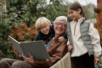 Senior Caucasian man sitting outdoors in village holding large book while smiling with two...