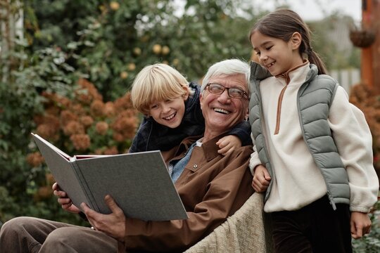 Senior Caucasian man sitting outdoors in village holding large book while smiling with two children, boy and girl, standing close and looking at book together - Powered by Adobe