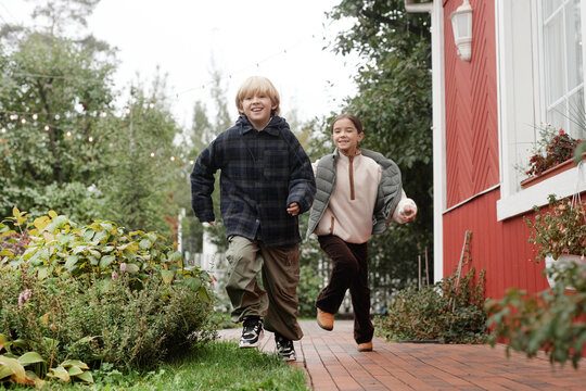 Boy and girl child running together along village path near red wooden house, smiling and looking forward, greenery and outdoor setting visible in background