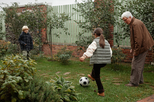 Senior Caucasian man watching two grandchildren playing soccer in village garden, girl kicking ball toward boy, outdoor family activity, greenery and wooden fence visible