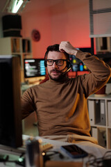Caucasian young adult man wearing headset sitting at desk working on computer in scam call center, raising hand to head with focused expression