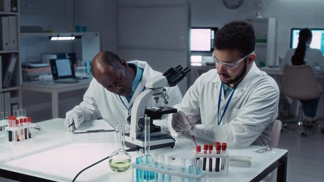 Medium panning shot of young polyethnic male student in white coat, goggles, gloves learning to perform blood test in microscope in bioengineering lab, guidance from senior African American supervisor