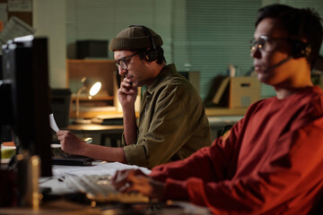 Biracial man wearing headset analyzing documents at computer while Indian man reading script inside...