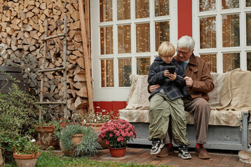 Senior Caucasian man sitting on bench in village holding smartphone with boy, both looking at...