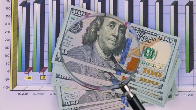 A stack of American dollars is examined through a magnifying glass. Slow motion.