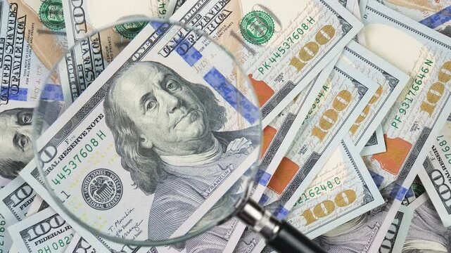 A stack of American dollars is examined through a magnifying glass. Slow motion.