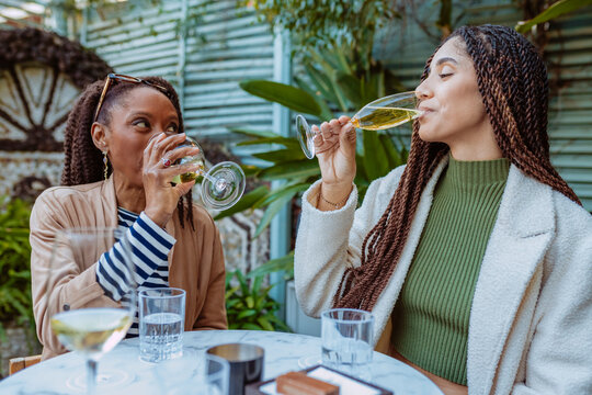 Multi ethnic mother and daughter sipping wine together at a stylish outdoor terrace.
