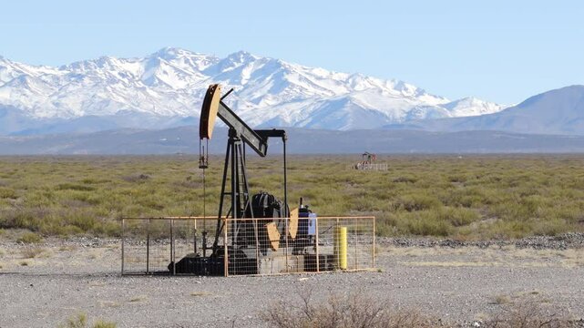Side drone shot of oil pumpjack in Vaca Muerta with snowy Andes mountains in the background.