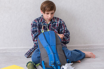 A young boy schoolboy sitting on the floor, unpacking his backpack at home. He is sad and boring.