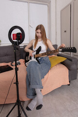 Young woman plays electric guitar on couch in cozy indoor setting while streaming a live session