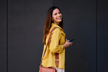 Smiling Woman Holding Smartphone While Wearing Casual Clothes Against Dark Wall