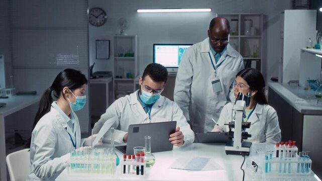 Full shot of mature African American bioengineering professor supervising group of polyethnic students learning to perform blood tests in laboratory, looking into microscope and filling report