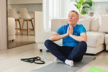 Selbstklebende Fototapeten Entspannung Yoga mindfulness meditation. Senior adult mature man practicing yoga at home. Mid age old grandfather sitting in lotus pose on yoga mat meditating relaxing. Older man doing breathing practice  © Юлия Завалишина