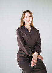 Portrait of a smiling young female doctor in dark uniform sitting on a stool with folded hands, isolated on white background.
