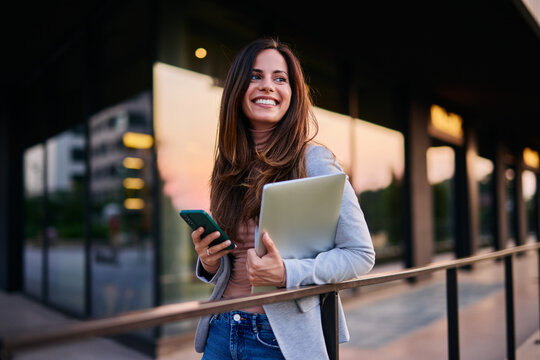 Smiling Professional Woman Holding Laptop and Phone in Modern Urban Setting
