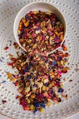 White plate with herbal tea from dried fruits and plants spilling out on a light background. A...