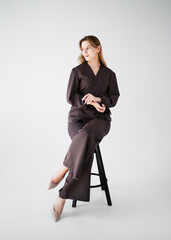 Portrait of a smiling young woman in a brown medical uniform sitting on a stool in studio on a light background. 