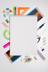 blank sketchbook lies in the center, surrounded by a variety of colorful stationery supplies, including markers, pencils, clips, and a protractor, arranged neatly on a white background