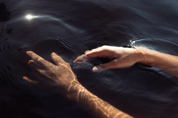 Fingers reach out to touch emerald waters, creating soft ripples as sunlight dances on the surface, adding a serene touch to the peaceful lakeside atmosphere at midday