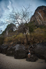 woman relaxes on a large rock along the sandy beach, with a towering cliff and a leafless tree in the background as dusk begins to settle over the serene coastal scene