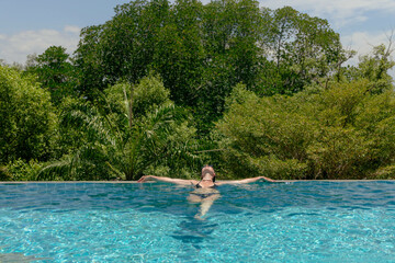 person enjoys leisure time by an infinity pool, stretching out while soaking in the sunlight. serene setting features vibrant green trees in the background, creating a peaceful ambiance
