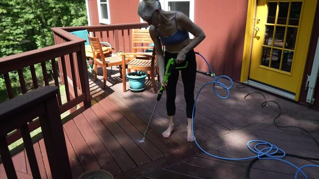 A mature woman utilizes a pressure washer to clean a wooden deck in her backyard, effectively removing dirt and stains from the surface.