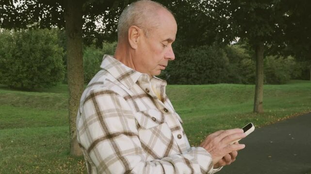 Positive elderly white man 65 years old enjoying technology, senior using modern smartphone, joyful moment surrounded by peaceful nature
