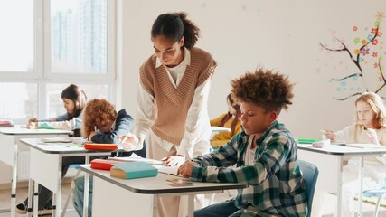 A schoolboy sits at a desk and talks and writes with a female teacher standing next to him while a group of four of his classmates sit next to them - Powered by Adobe
