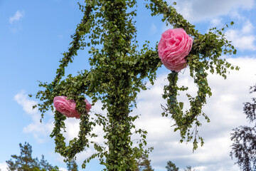 A Swedish midsummer pole decorated with green birch leaves and pink paper flowers against a partly...