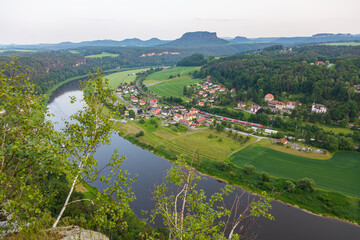 Scenic view over rural village of Rathen in Saxon Switzerland, Germany, with a red train passing through the Elbe valley, surrounded by forested hills, farmland, and sandstone cliffs in summer light.
