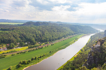 Scenic panoramic view from the Bastei cliffs over the Elbe River valley with sandstone rocks, lush forests, rural village rooftops, distant mountain ranges under a vibrant summer sky. Saxony, Germany