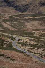A solitary figure traverses the rugged, winding path through the majestic and mist-shrouded peaks of the Scottish Highlands, possibly in the iconic Glen Coe, where ancient landscapes meet dramatic ski