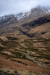 A solitary figure traverses the rugged, winding path through the majestic and mist-shrouded peaks of the Scottish Highlands, possibly in the iconic Glen Coe, where ancient landscapes meet dramatic ski