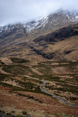 A solitary figure traverses the rugged, winding path through the majestic and mist-shrouded peaks of the Scottish Highlands, possibly in the iconic Glen Coe, where ancient landscapes meet dramatic ski