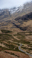 A solitary figure traverses the rugged, winding path through the majestic and mist-shrouded peaks of the Scottish Highlands, possibly in the iconic Glen Coe, where ancient landscapes meet dramatic ski
