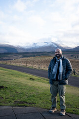 A guy in a scenic view of a road leading into a vast, rugged landscape with snow-capped mountains under a cloudy sky, likely in the Scottish Highlands