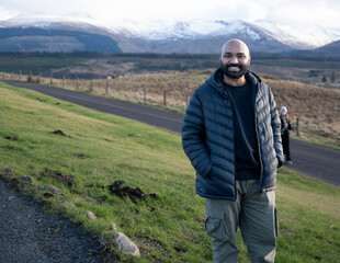 A guy in a scenic view of a road leading into a vast, rugged landscape with snow-capped mountains under a cloudy sky, likely in the Scottish Highlands