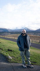 A guy in a scenic view of a road leading into a vast, rugged landscape with snow-capped mountains under a cloudy sky, likely in the Scottish Highlands